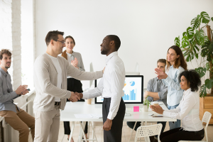 Young black man getting promoted and shaking hands with white men in front of his co-workers