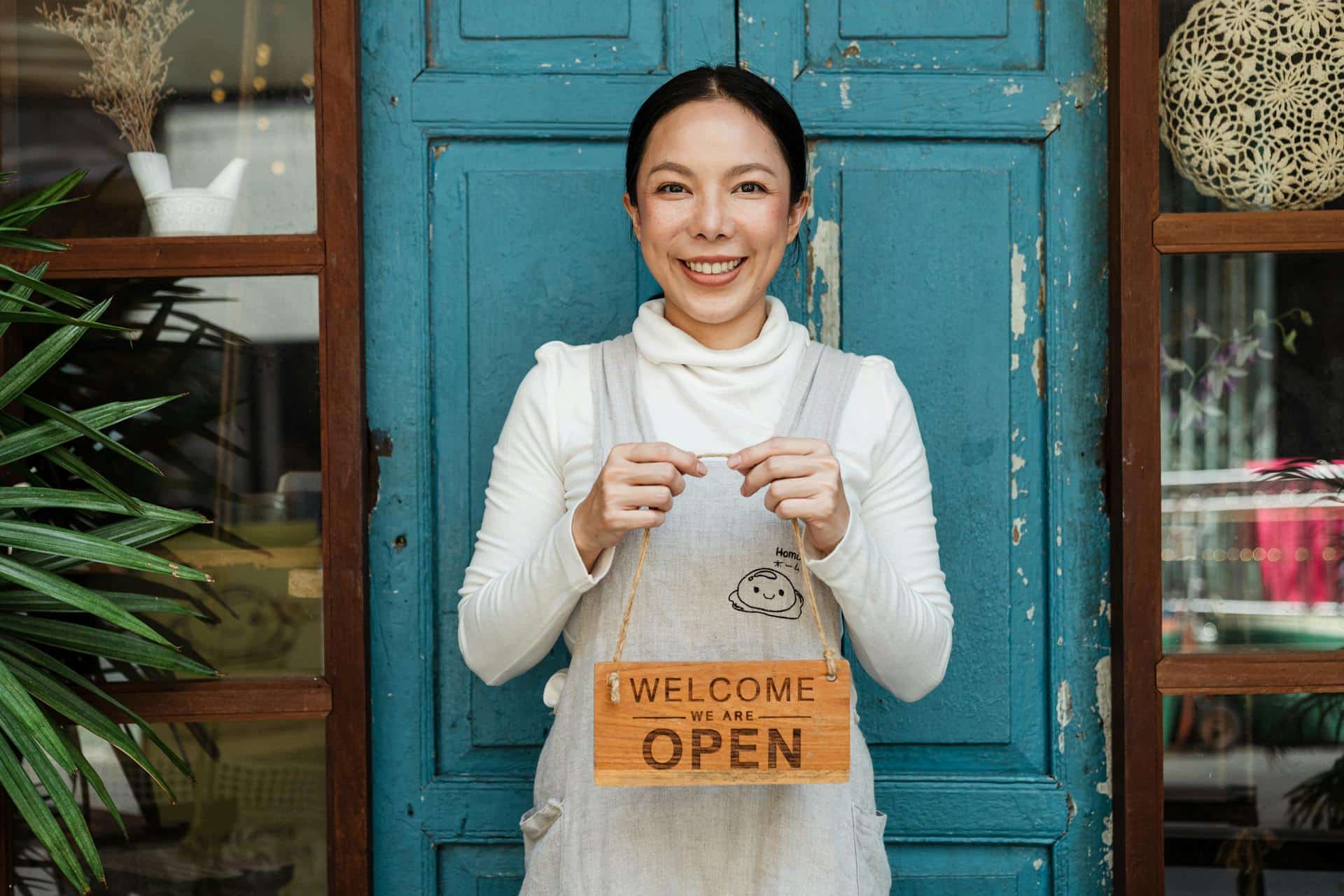 “a shop owner standing in front of a door holding a sign that says WELCOME WE ARE OPEN