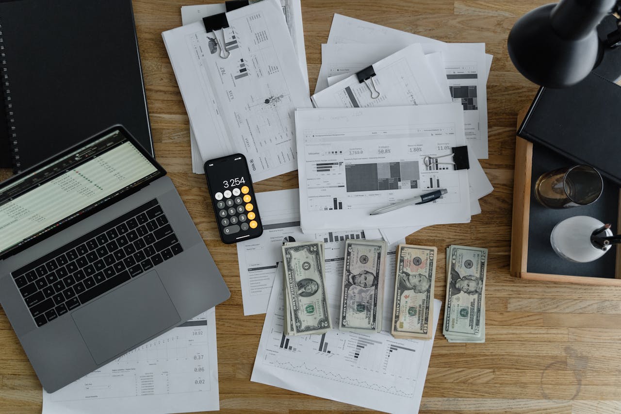 A wooden desk with a laptop, phone, papers, and money